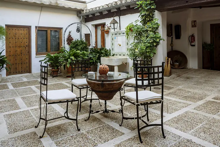 A courtyard under a timber-beamed loggia: a stone table, climbing plants and a pebbled floor, opening into the house.