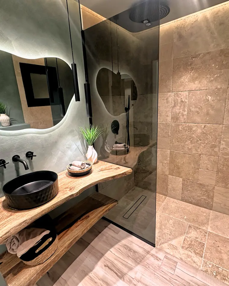 Bathroom in JLT: stone basin on a live-edge timber shelf, organic backlit mirror, travertine walls.