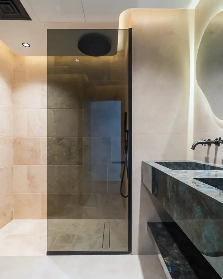 Bathroom in Dubai Marina: travertine walk-in shower, a monolithic dark stone basin and a backlit organic mirror.