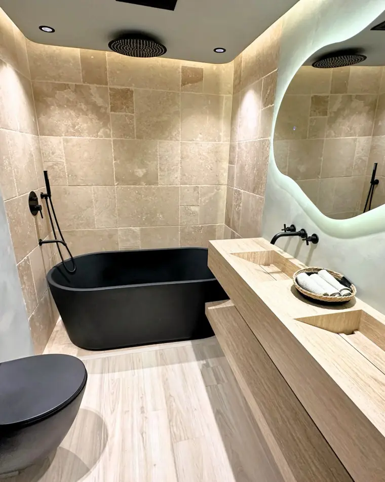 Bathroom in TECOM: black freestanding bath, travertine walls and a backlit organic mirror.