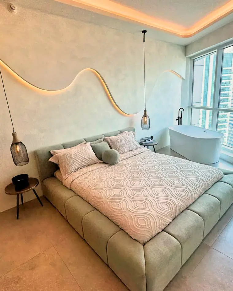 Bedroom in JLT: olive upholstered bed, a serpentine cove light, a freestanding bath and a city view.