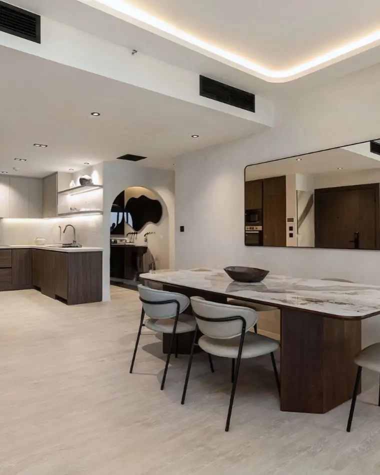 Dining room in JBR: a serpentine cove ceiling, a marble table and pale oak flooring, with the kitchen beyond.