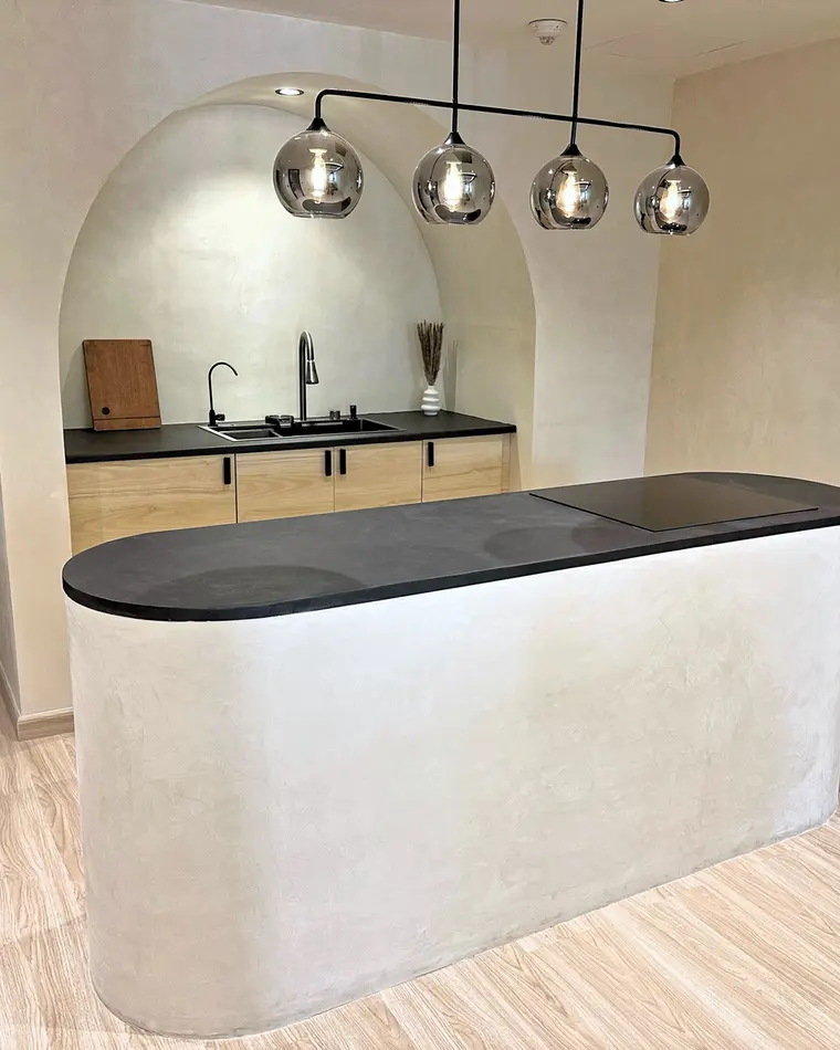Kitchen in JBR: microcement arch, curved island with a dark stone top, four smoked-glass pendants.