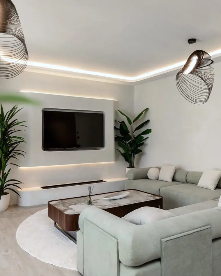 Living room in JBR: sage green corner sofa, olive trees, cove lighting and a wall-mounted television.