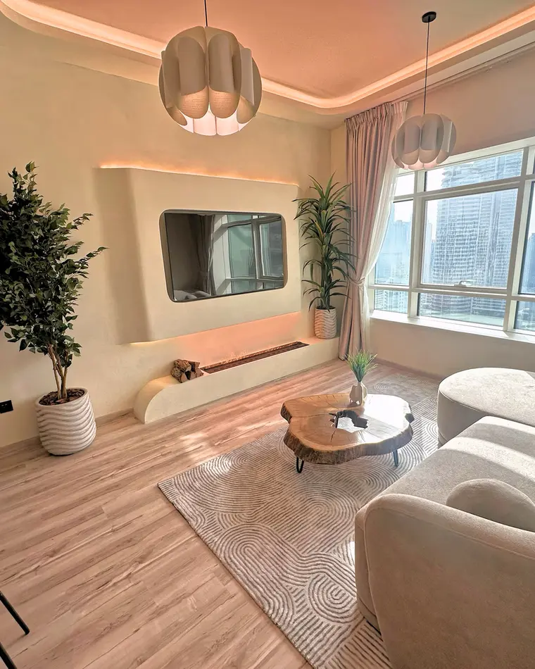 Living room in JLT: sectional sofa facing a wall-mounted television over a linear fireplace, with a rattan pendant.